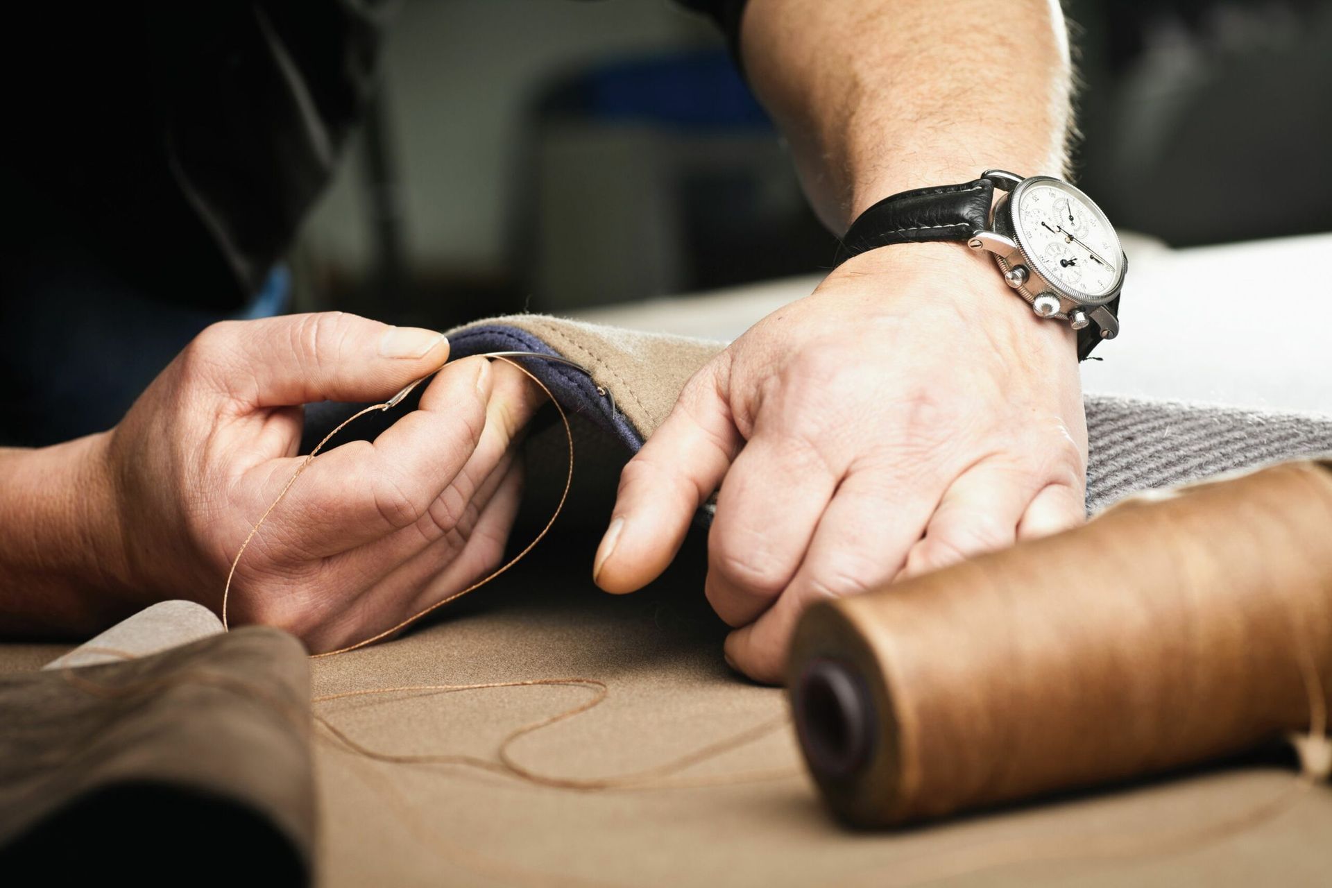 Personenhände nähen Leder mit Nadel und Faden; eine Garnrolle ist sichtbar.