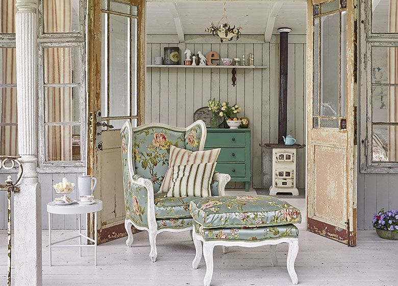 Veranda im Shabby-Chic-Stil mit geblümtem Sessel und Hocker, türkisfarbener Kommode und Holzofen.
