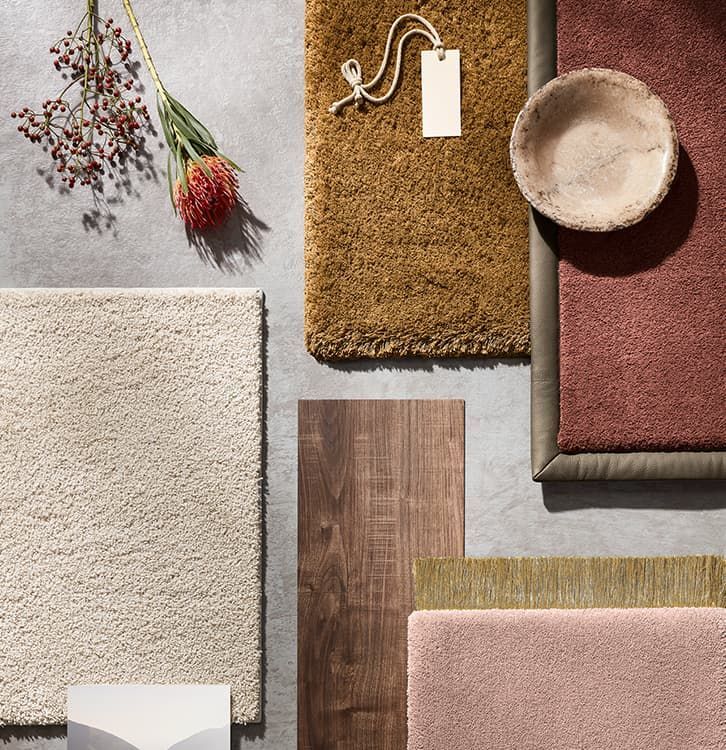 Draufsicht auf verschiedene Texturen: Teppiche, Holz, Schale, Blume, Etikett. Erdtöne vor grauem Hintergrund.