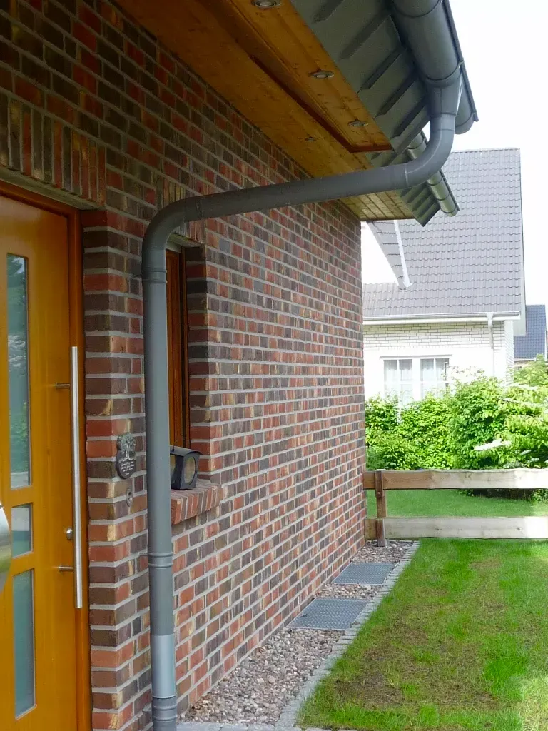 Ein graues Metallfallrohr verläuft entlang einer Ziegelmauer neben einer Holztür und führt vom Dachüberstand zum Boden.