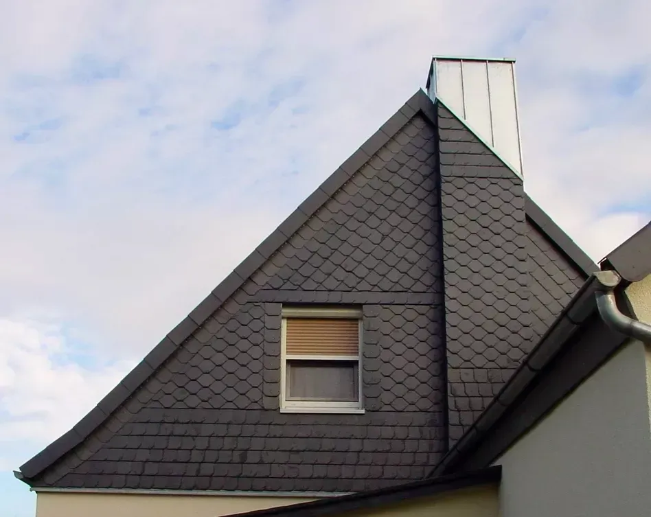 Eine steile, dunkelgrau geflieste Giebelwand mit einem einzelnen quadratischen Fenster mit geschlossenen Jalousien und einem darüber liegenden metallenen Schornstein.