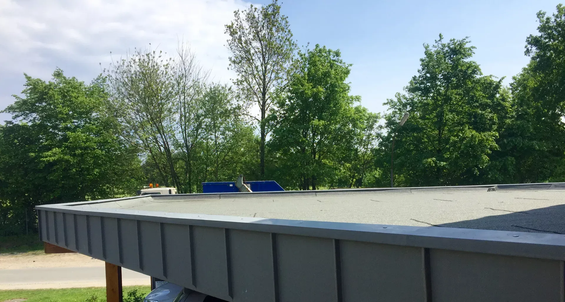 Eine flache Dachkante mit grauen Metallleisten vor dem Hintergrund grüner Bäume unter strahlend blauem Himmel.
