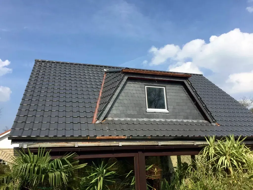 Ein dunkel geziegeltes Hausdach mit Dachgaube vor blauem Himmel mit sanften weißen Wolken und grünem Laub darunter.