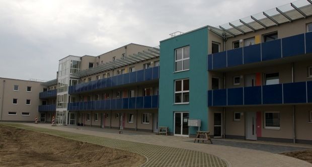 Ein dreistöckiges, modernes Apartmentgebäude mit beigen Wänden, blauen Balkonen und einem markanten türkisfarbenen Bereich.