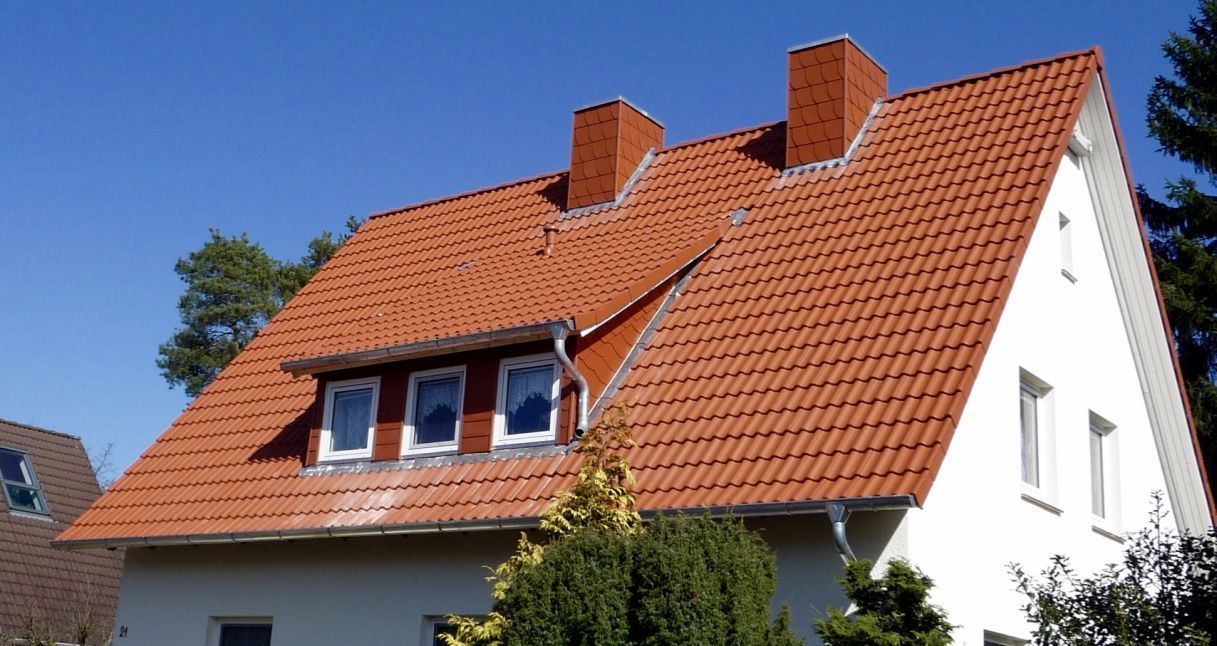 Ein zweistöckiges Haus mit weißer Fassade und steilem, rot gedecktem Dach mit Dachgaube und zwei gemauerten Schornsteinen.