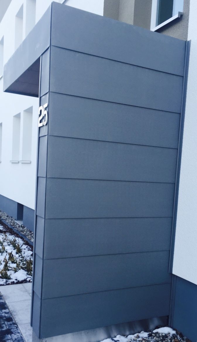 Ein modernes, graues Metallvordach am Eingang und Fassadenverkleidung an einem Gebäude mit der Hausnummer 25 an der Ecke.
