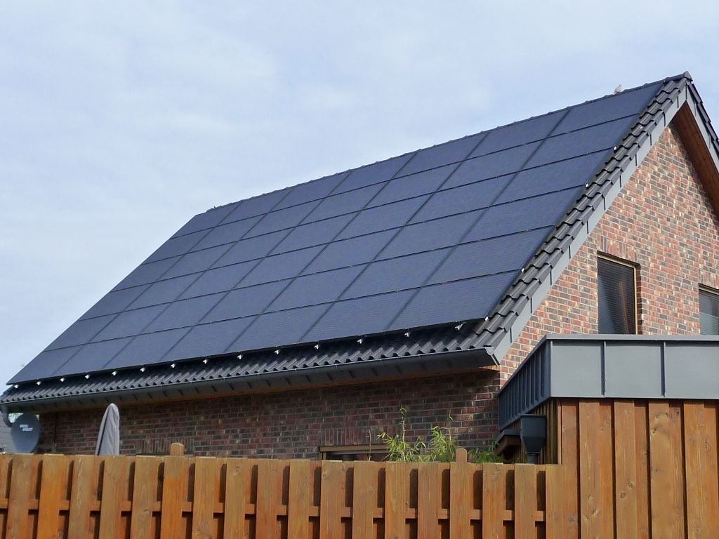 Ein Backsteinhaus mit einem dunklen, vollflächig mit Solarpaneelen bedeckten Dach, gesehen von hinter einem Holzzaun.