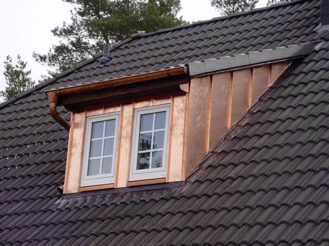 Ein kupferverkleidetes Dachgaubenfenster in einem dunkelgrauen Ziegeldach vor dem Hintergrund von Kiefern.