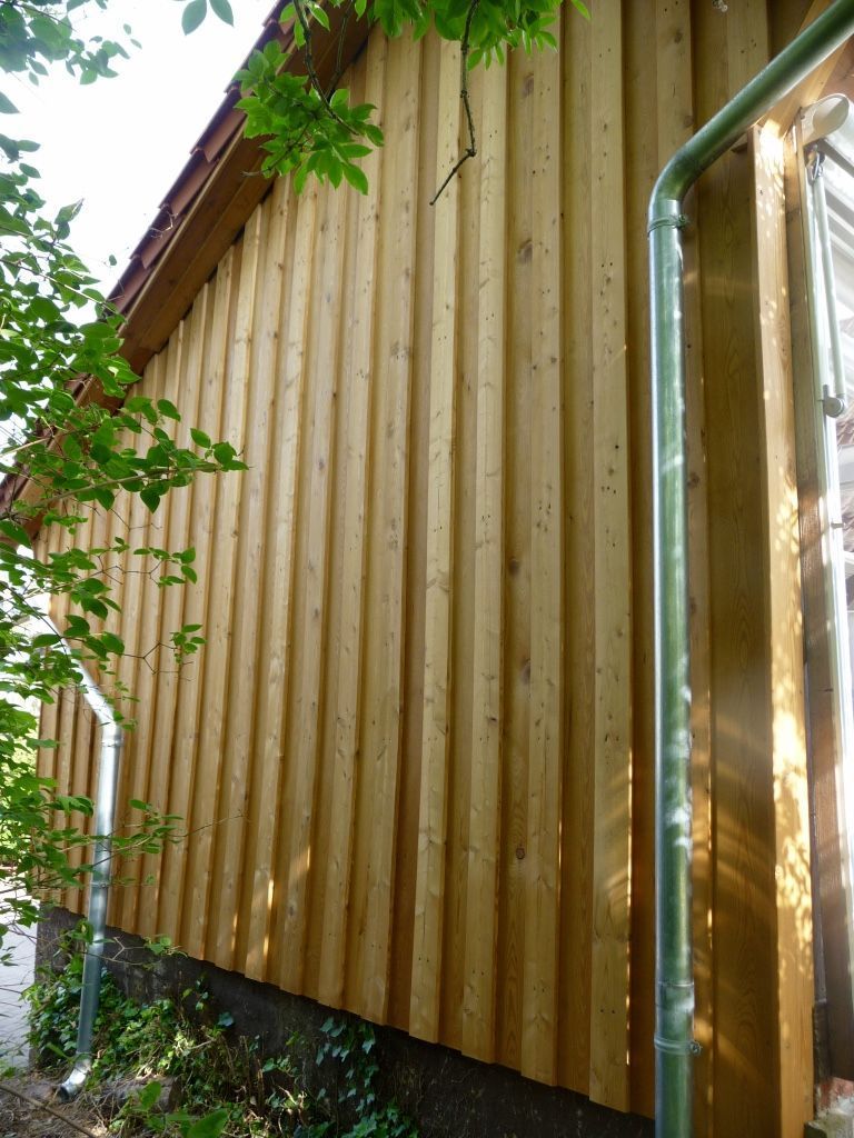 Seitenansicht einer Gebäudeaußenfassade mit heller, vertikaler Holzverkleidung in Stülpschalung und einem Fallrohr aus Metall.