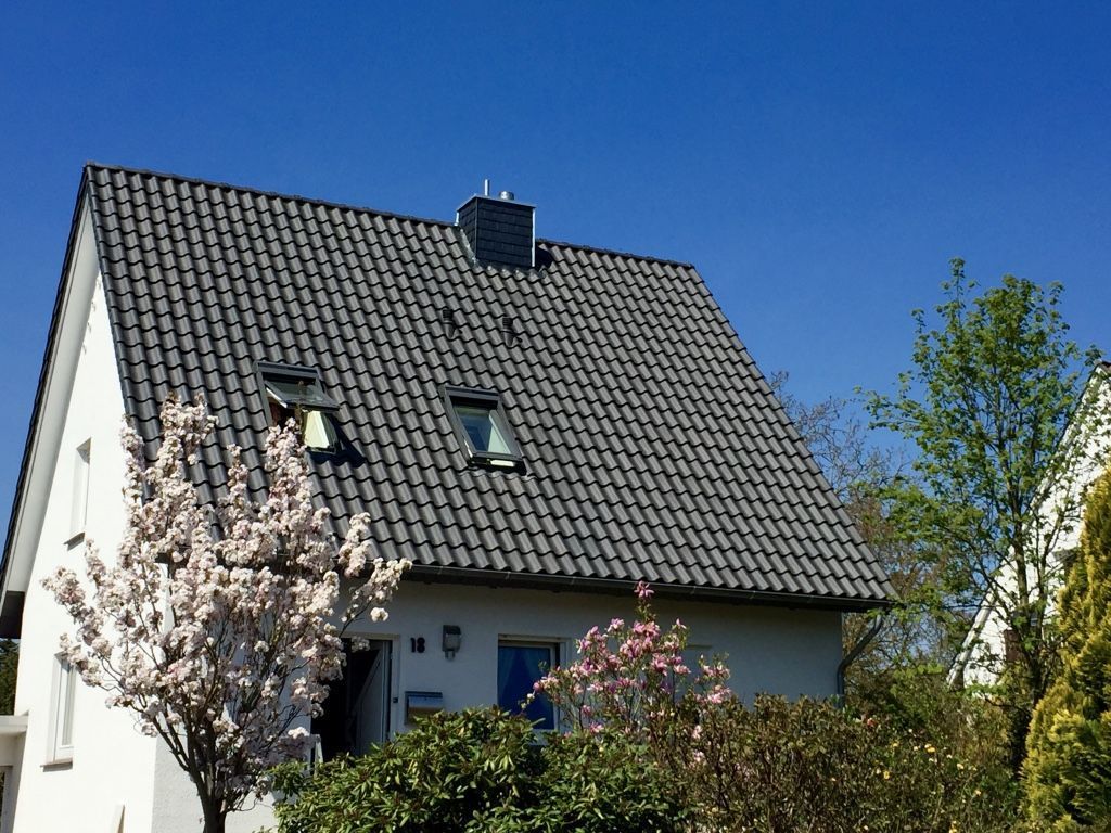 Ein weißes Haus mit dunklem Giebeldach und zwei Dachfenstern vor klarem, blauem Himmel, davor ein blühender Baum.