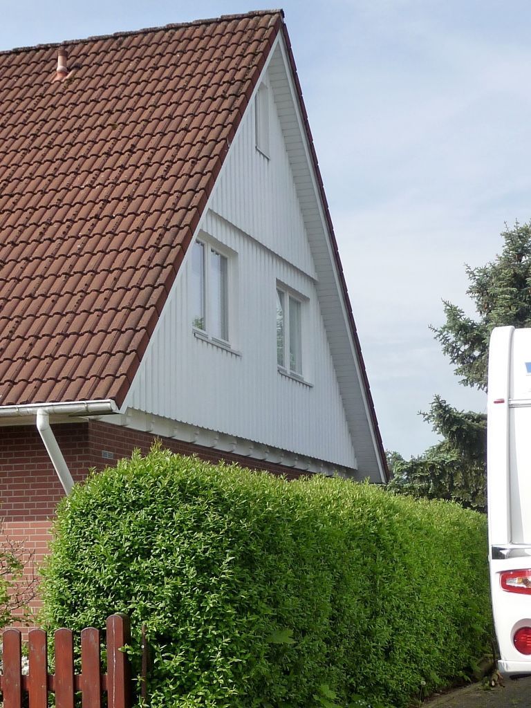 Hinter einer hohen, grünen Hecke steht unter klarem Himmel ein weißes Giebelhaus mit rotem Ziegeldach.