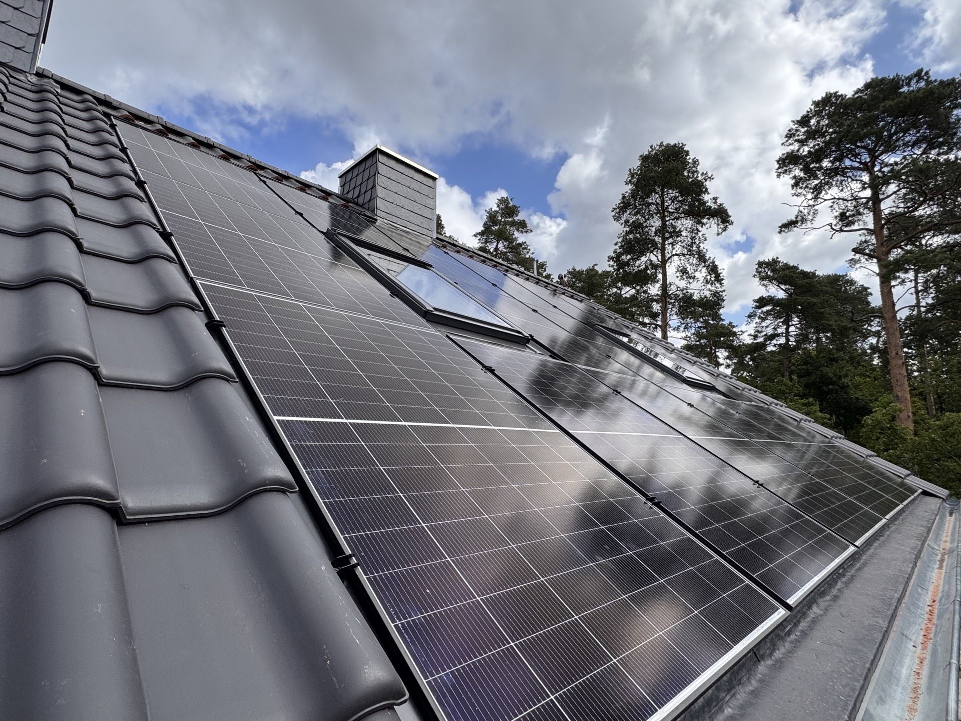 Auf einem dunklen Ziegeldach installierte Solarpaneele vor blauem Himmel mit Wolken und Bäumen im Hintergrund.