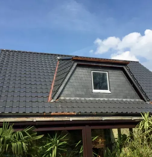 Ein dunkel gedecktes Ziegeldach mit Dachgaube, von unten gesehen vor einem blauen Himmel mit leichten Wolken.