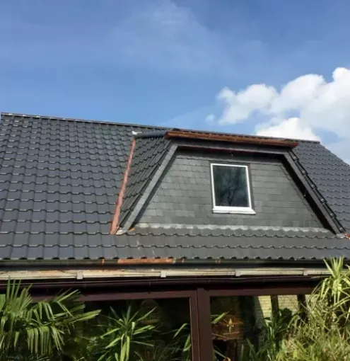 Ein dunkel gedecktes Ziegeldach mit Dachgaube, von unten gesehen vor einem blauen Himmel mit leichten Wolken.