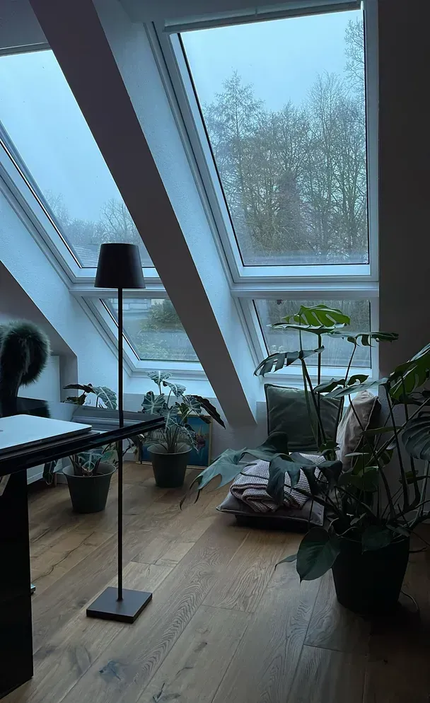 Dachzimmer mit schrägen Dachfenstern, Holzfußboden, einer hohen schwarzen Stehlampe und mehreren Topfpflanzen.