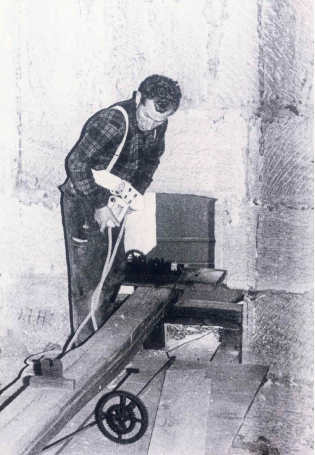 Schwarzweißbild eines Mannes mit lockigem Haar und kariertem Hemd, der mit gesenktem Blick an einer rauen Wand arbeitet.
