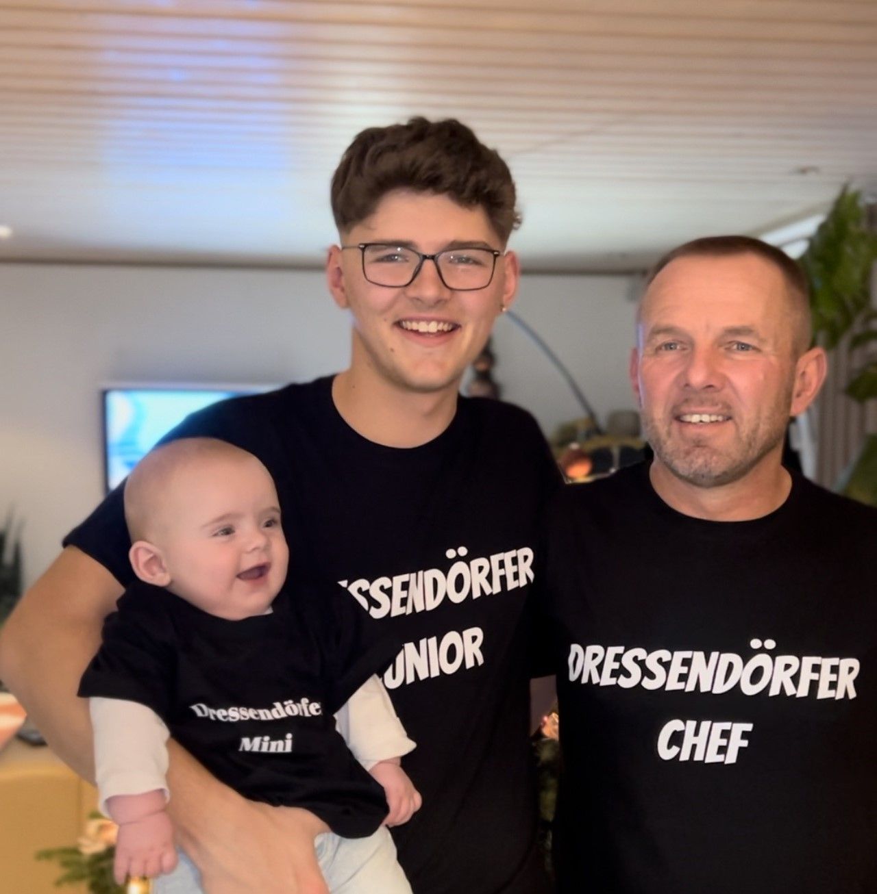 Ein junger Mann mit Brille steht lächelnd neben einem älteren Mann; im Vordergrund ist teilweise ein Baby zu sehen.
