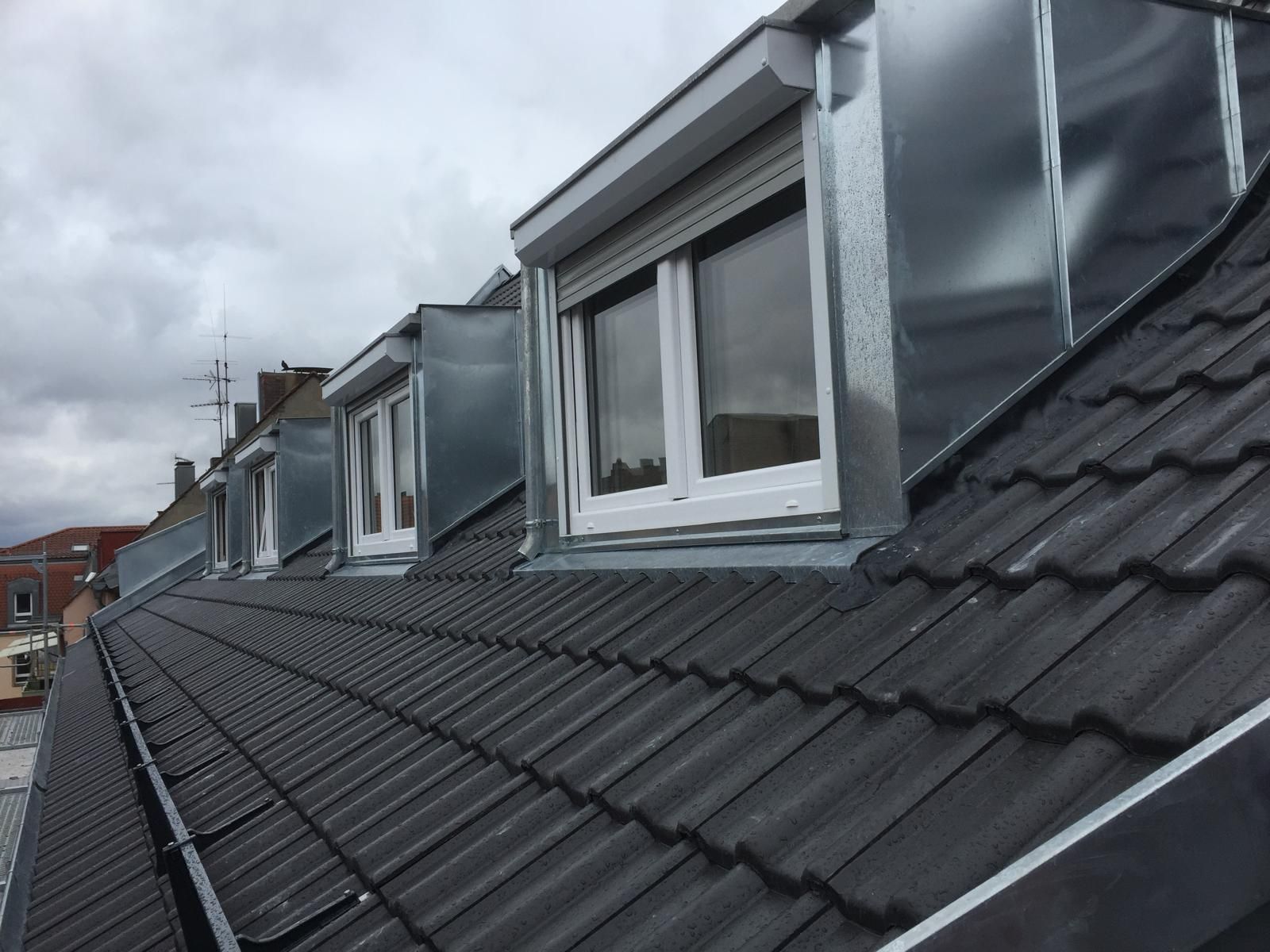 Steildach mit Betondachsteinen, mehrfachen Gaubenaufbauten, Fensteranlagen und Metallverkleidung entlang der Dachfläche