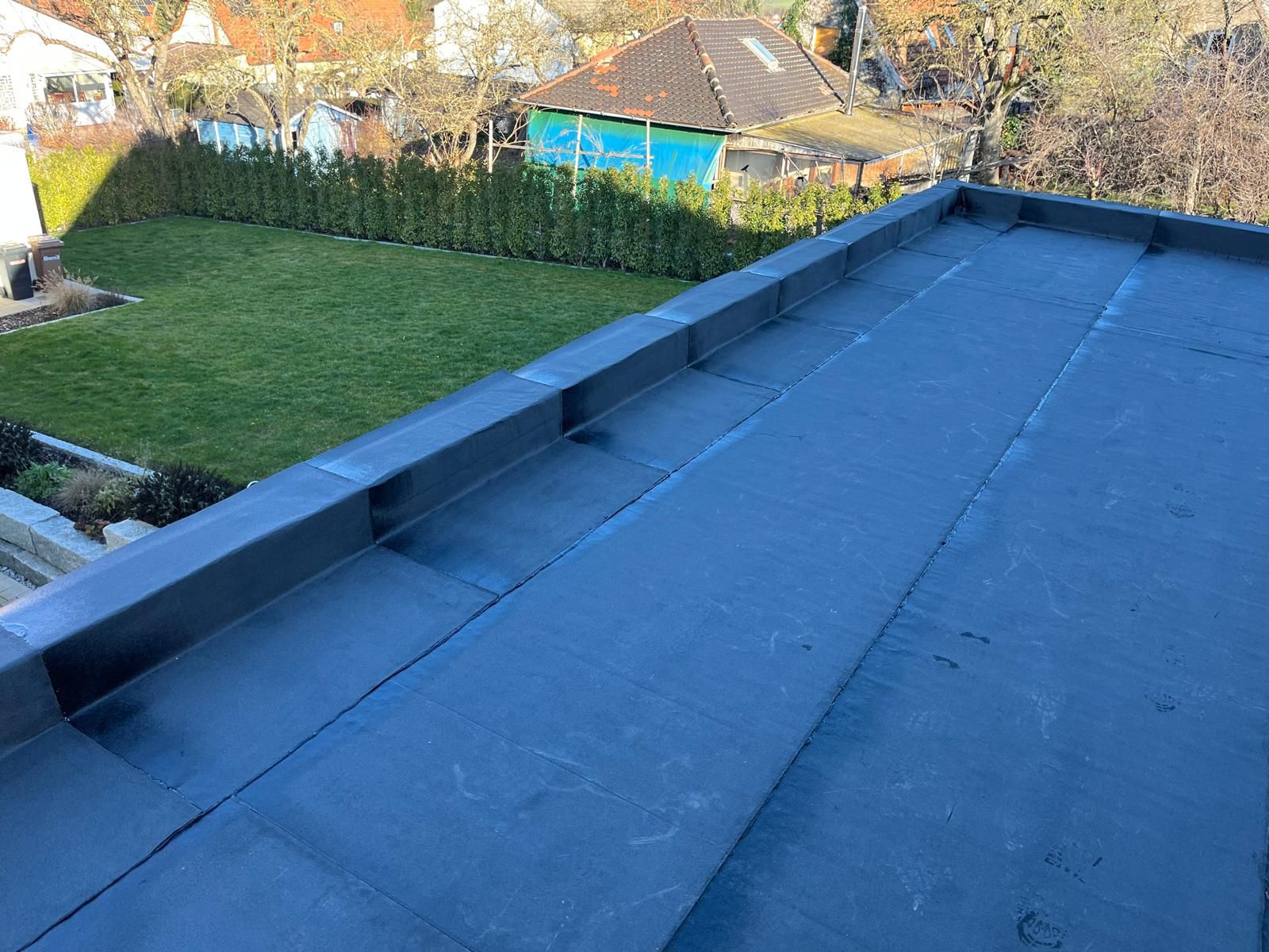 Flachdach mit dunkler Bitumenabdichtung und erhöhtem Rand, im Hintergrund ein Garten mit Rasenfläche