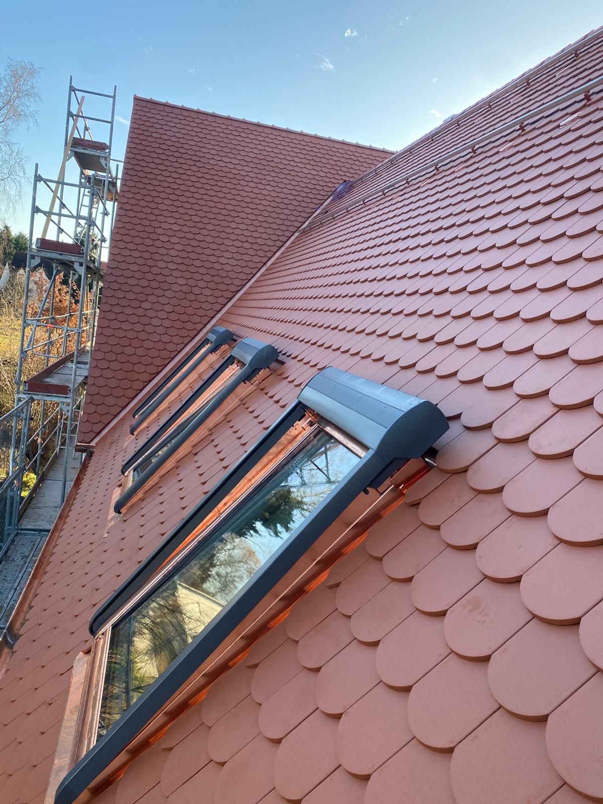 Rotes Ziegeldach mit eingebautem Dachfenster während einer Dachsanierung, daneben ein Baugerüst unter blauem Himmel.