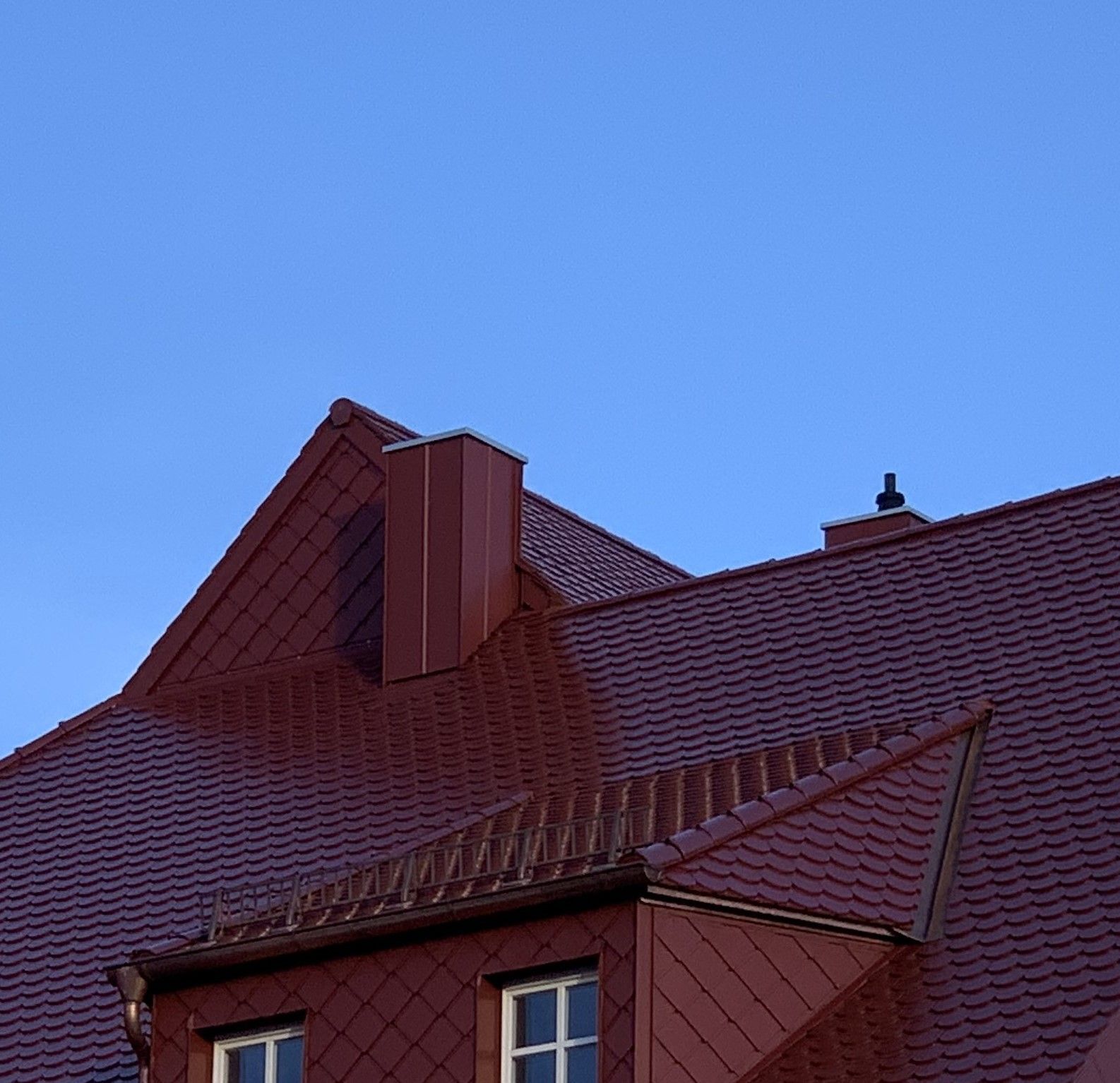 Zwei kupferfarbene Kamine ragen aus dunkelrotem Ziegeldach vor klarem blauem Himmel