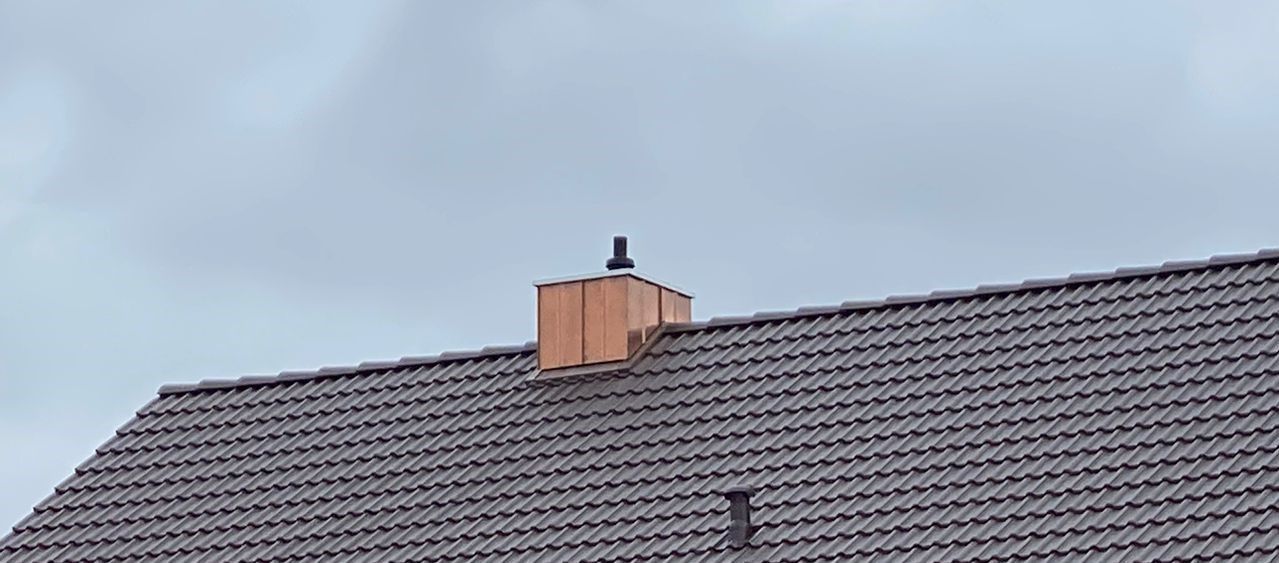 Neuer verkleideter Kaminkopf mittig auf dunkelgrauem Ziegeldach vor bewölktem Himmel