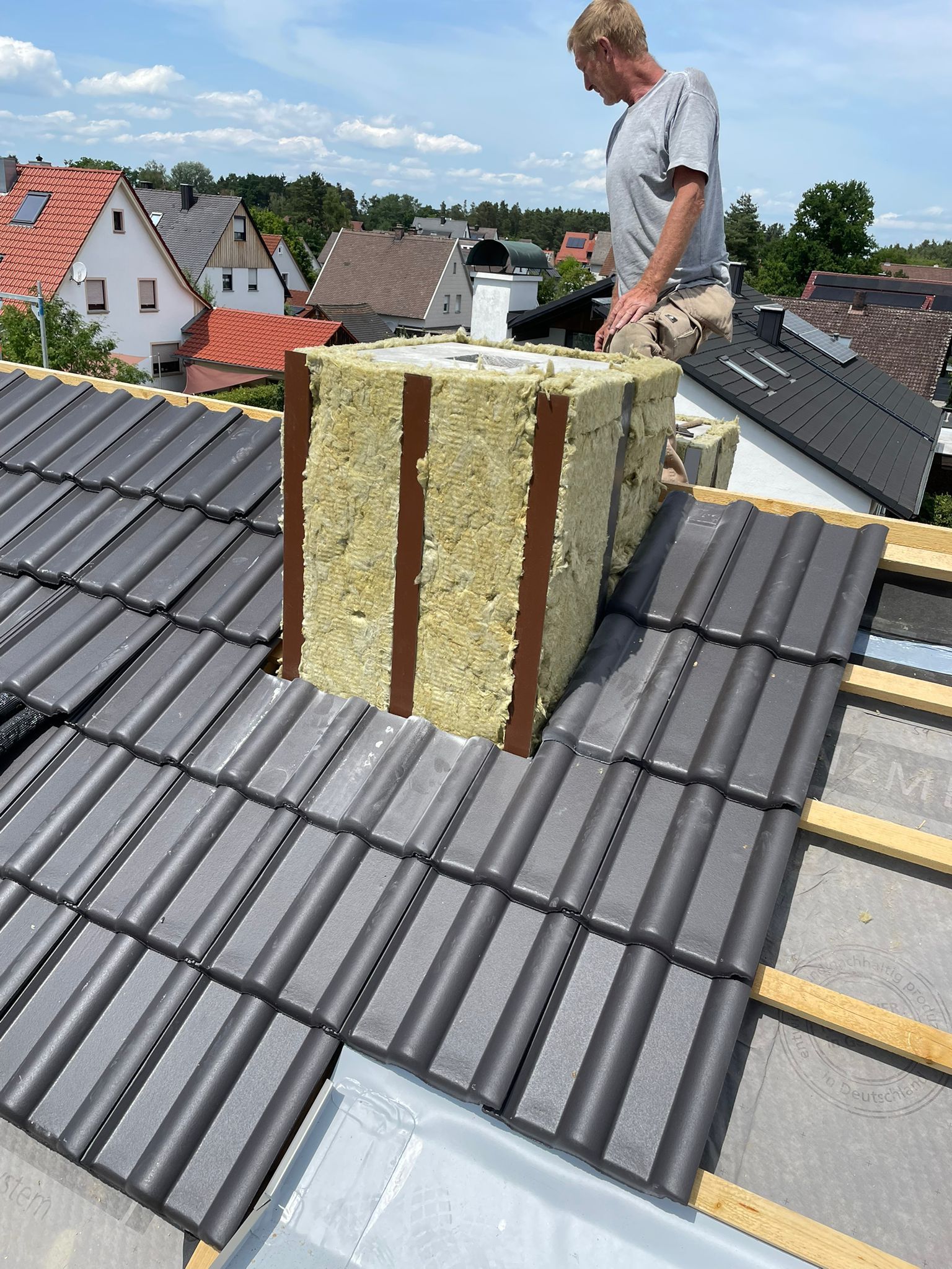 Handwerker auf einem Hausdach installiert Dämmerung aus Mineralwollean einem Schornstein, im Hintergrund sind Wohnhäuser.