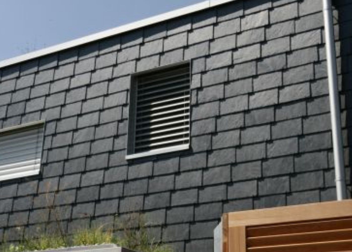 Hausfassade mit dunkelgrauen, rechteckigen Schieferplatten in waagerechter Deckung; mittig ein Fenster mit Lamellenjalousie.