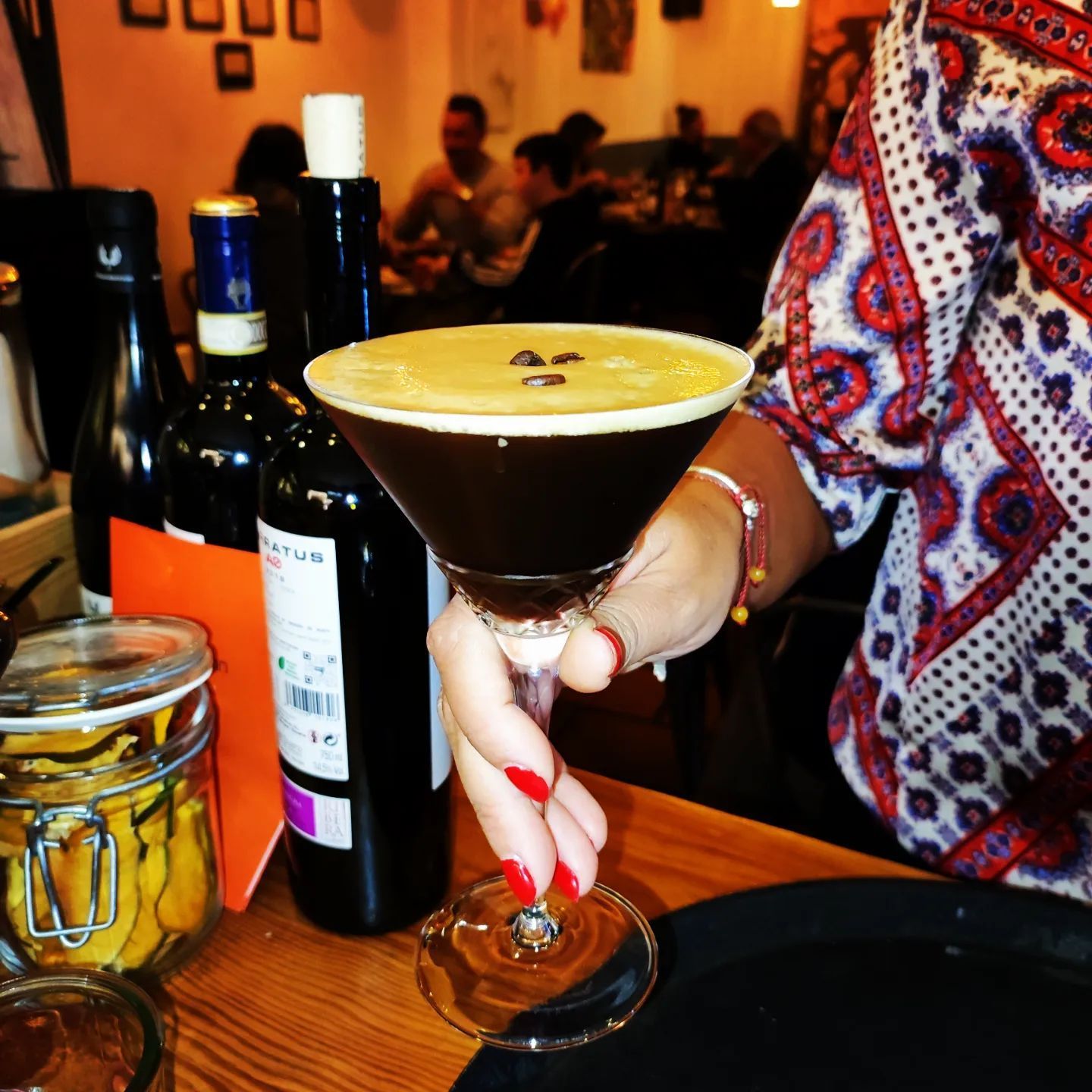 Una mujer sostiene un martini con una botella de vino al fondo.
