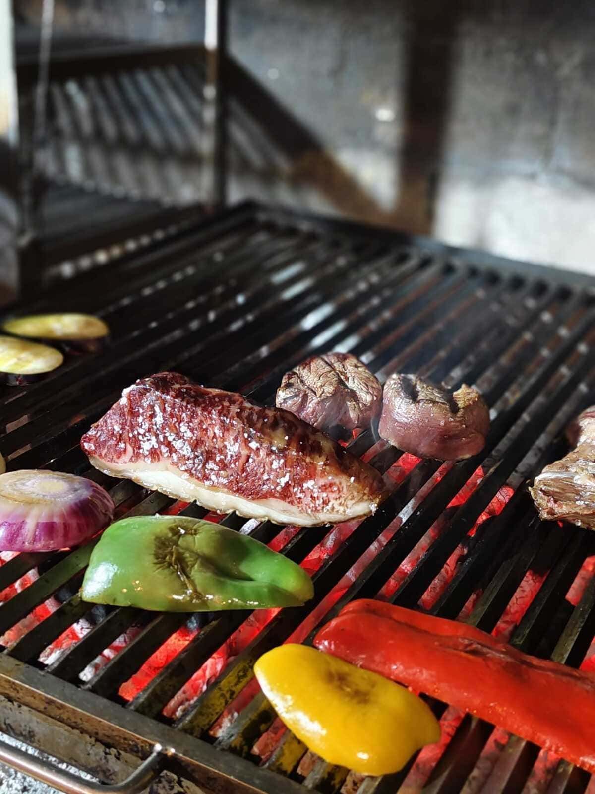 Verduras y un filete asándose a la parrilla, con brasas al rojo vivo y humo visible.