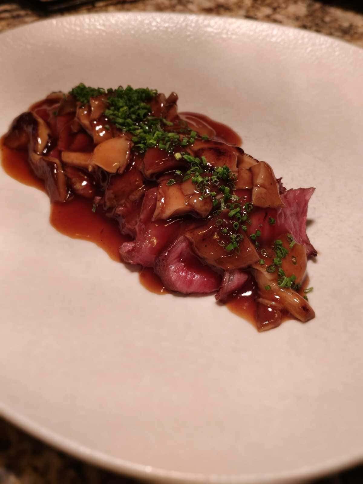 Filete en rodajas cubierto con champiñones y hierbas en un plato blanco.