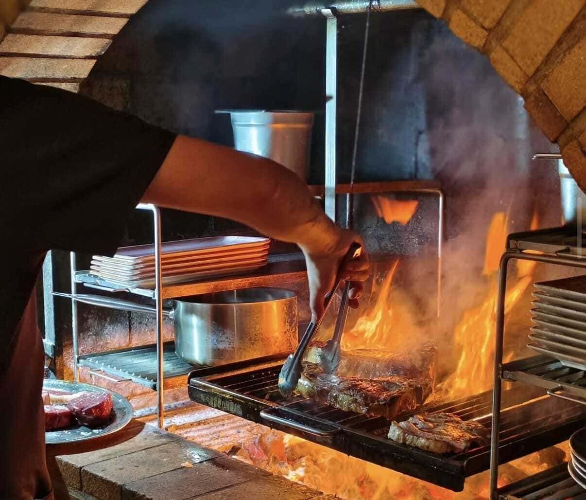 Persona asando carne en un horno de leña, con llamas y humo en una cocina de ladrillo.