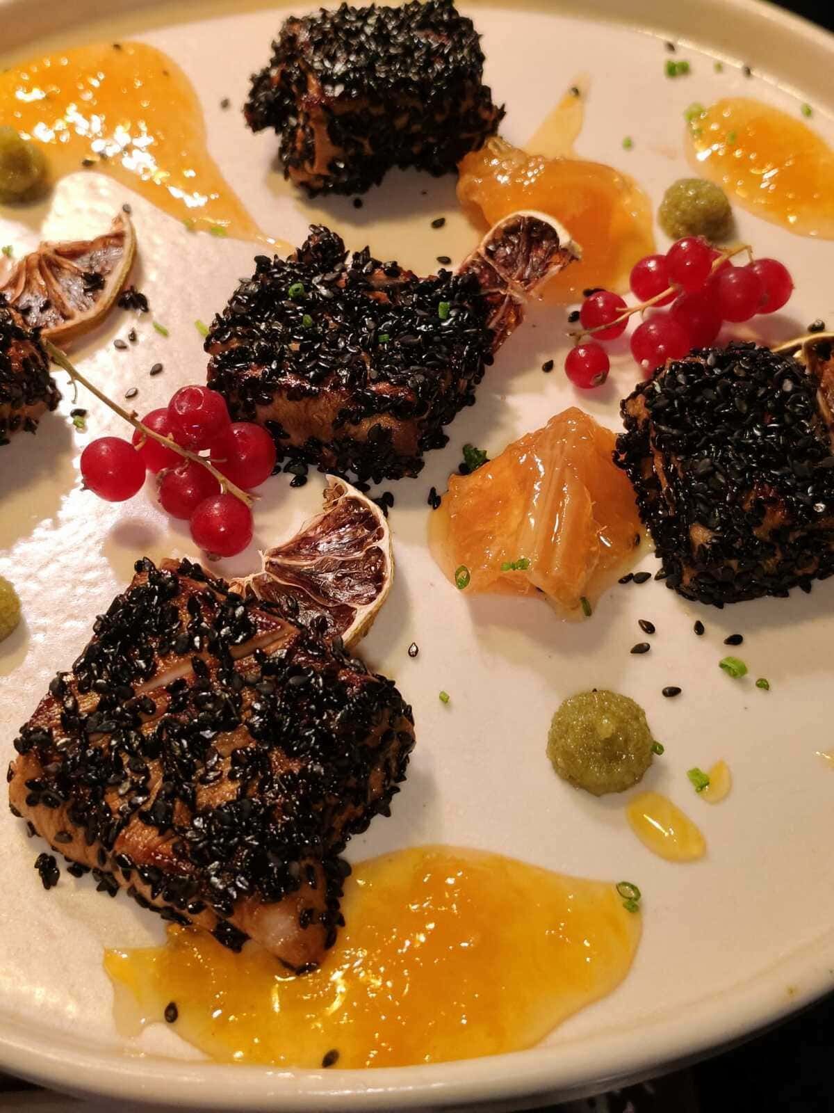 Elegante postre emplatado con trozos de migas oscuras, gajos de naranja, grosellas rojas y flores comestibles sobre un plato blanco.