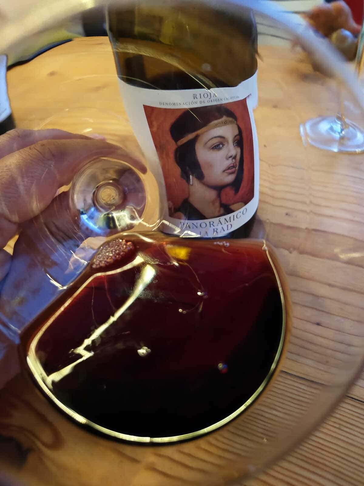 Una copa de vino tinto junto a una botella con una etiqueta que muestra un retrato, sobre una mesa de madera.