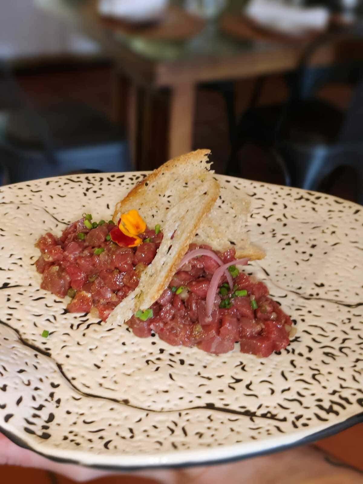 Dos tortillas sobre un plato moteado, cubiertas con atún crudo picado, cebolla y flores comestibles.