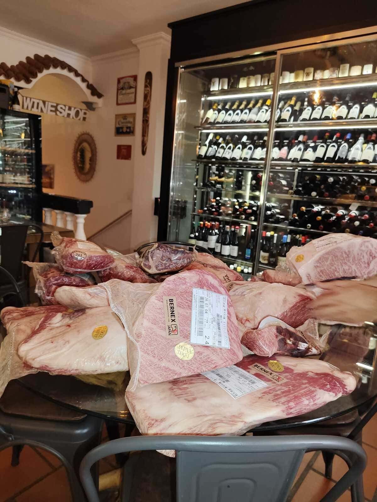 Mesa repleta de grandes jamones curados de color rosa en la bodega de una vinoteca, con botellas de vino alineadas en la pared.