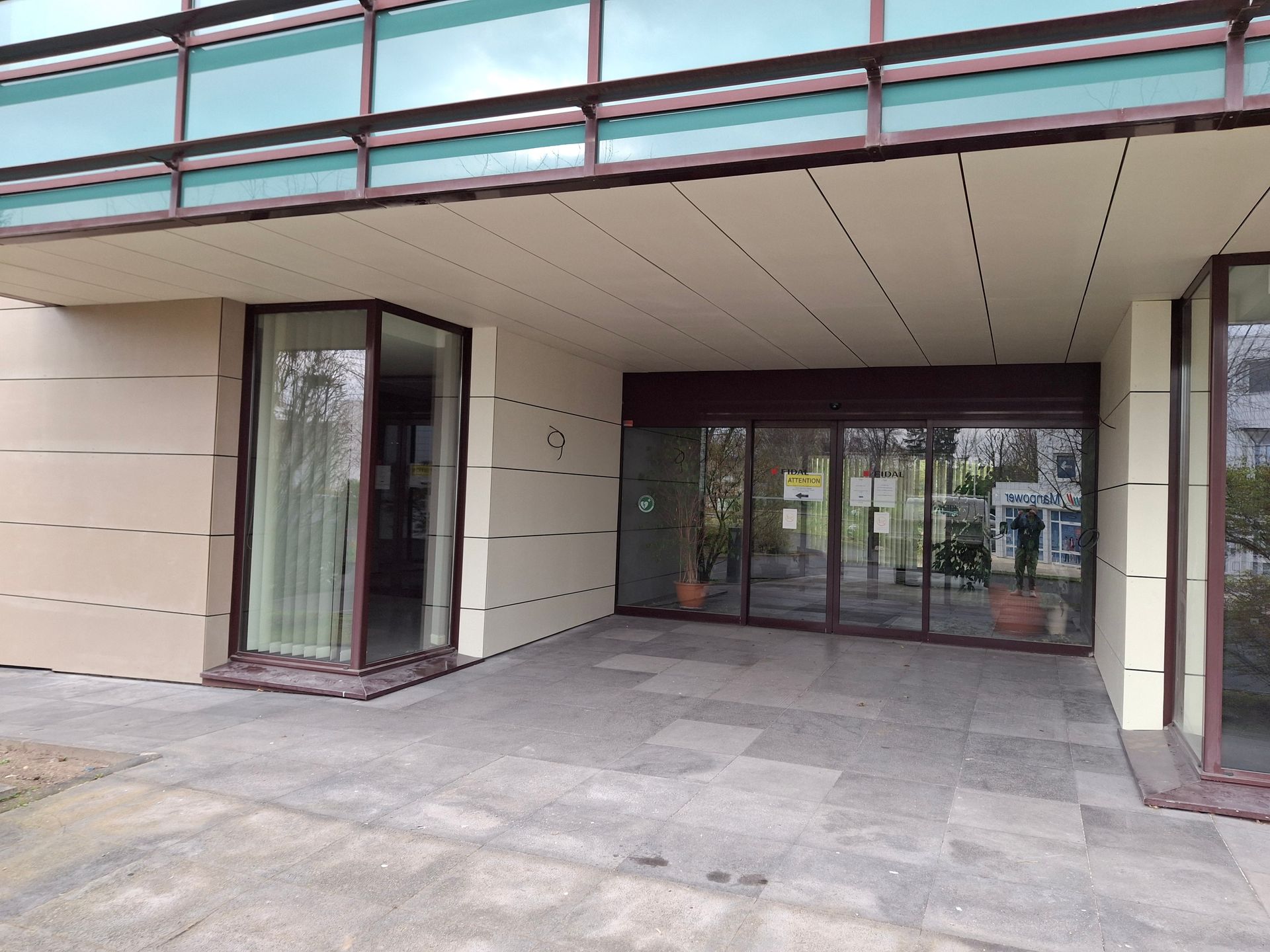 Entrée de l'immeuble avec portes et fenêtres vitrées, murs beige clair et allée carrelée.