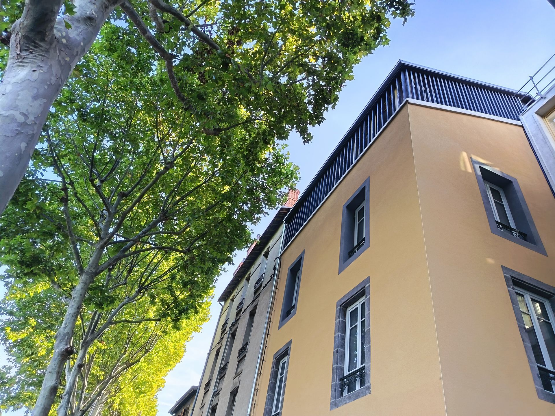 Bâtiment jaune au toit sombre et entouré d'arbres, vu d'en bas. Ciel bleu au-dessus.