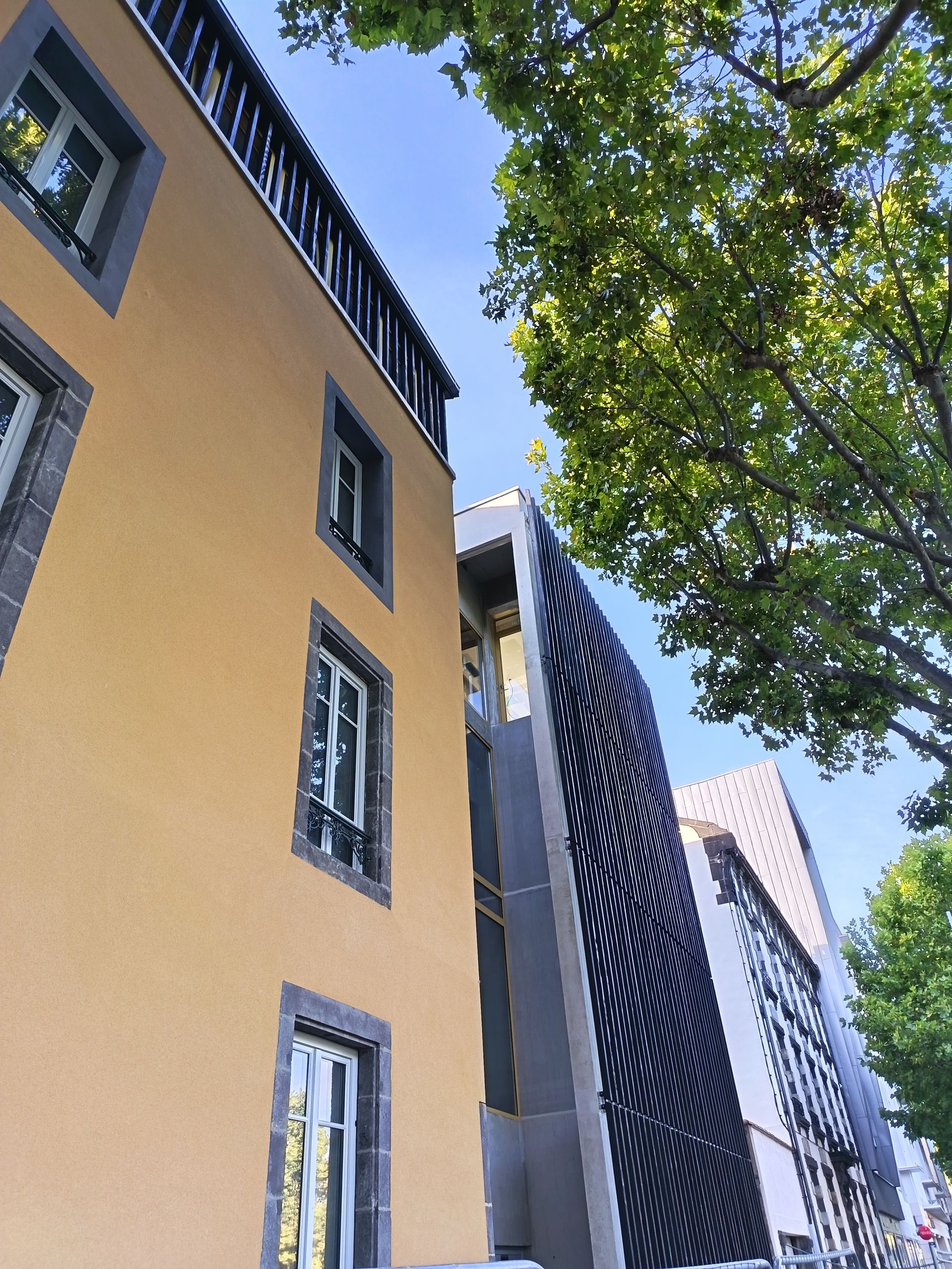 Bâtiments aux façades de styles variés : jaunes, modernes avec un écran noir incurvé, et blanches. Ciel bleu.