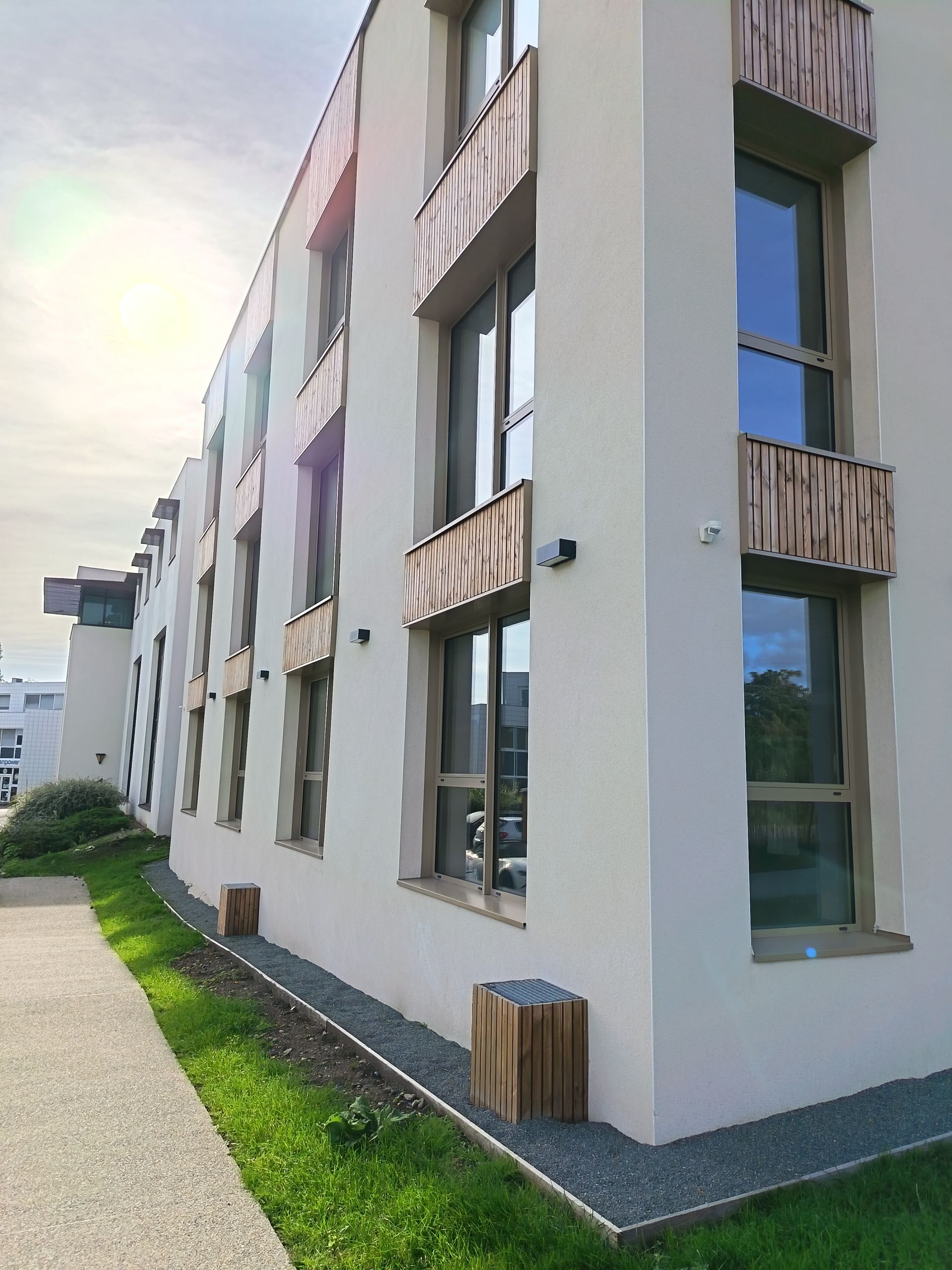 Bâtiment moderne à façade blanche et éléments en bois autour des fenêtres. Une allée et un espace vert au premier plan.