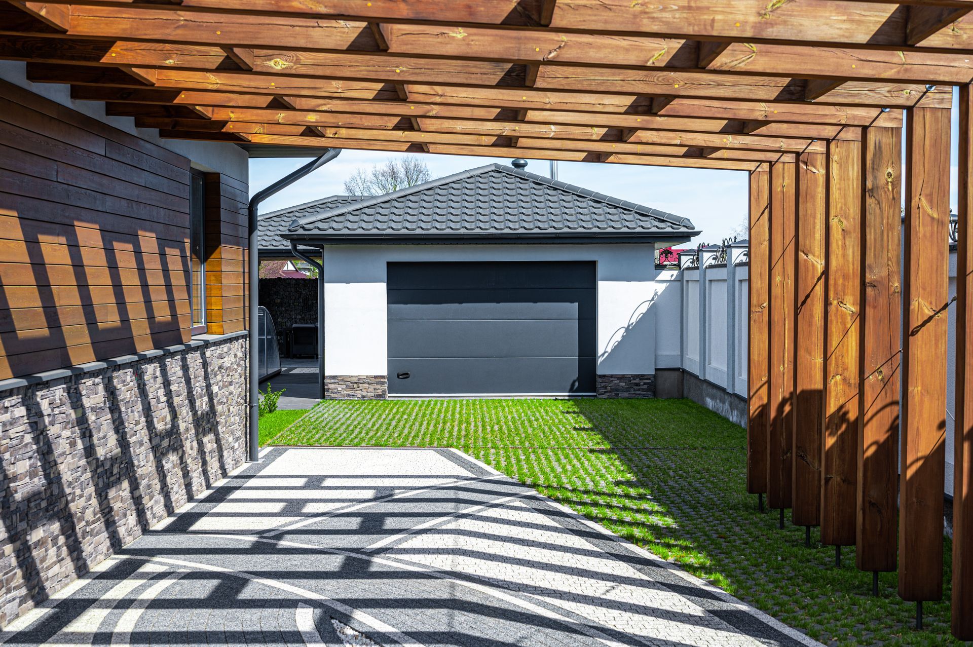 Une pergola en bois encadre une allée menant à un garage doté d'une porte grise. Pelouse verte et pavés.
