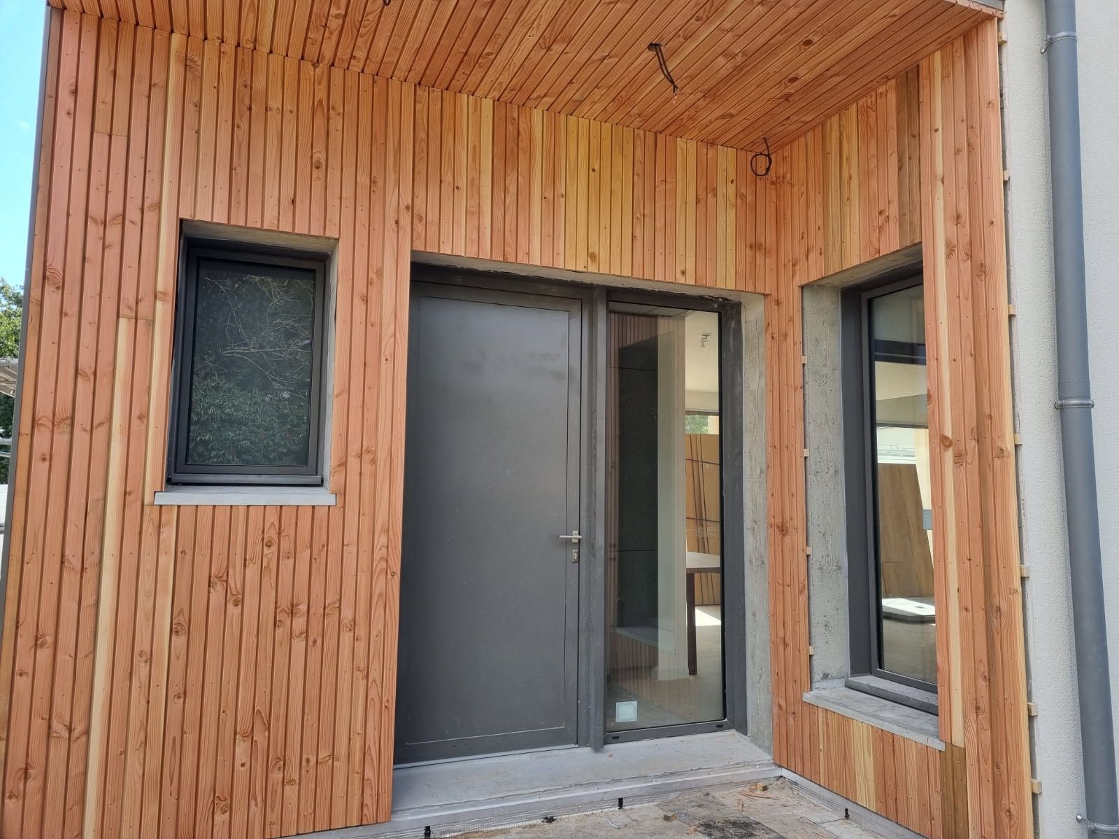 Entrée revêtue de bois avec une porte grise et deux fenêtres à cadre sombre.