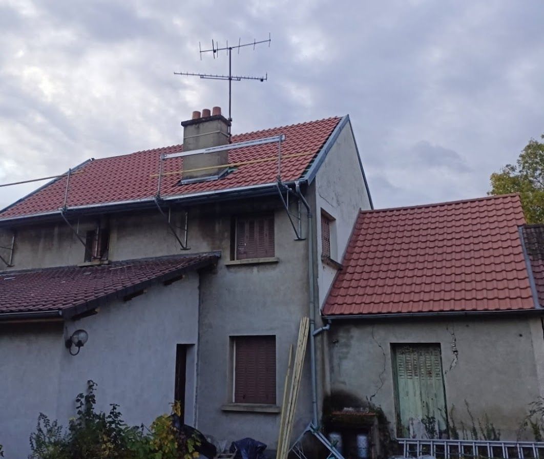 Maison à étages, avec toit de tuiles rouges, cheminée et antenne, sous un ciel nuageux.