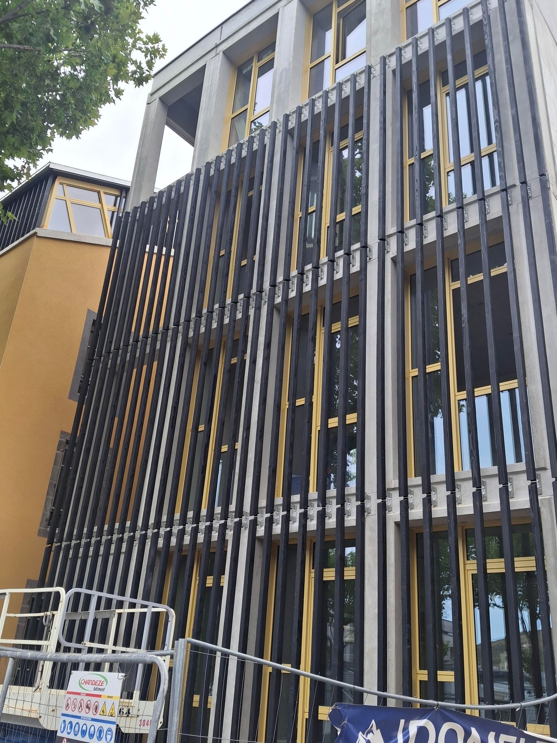 Bâtiment moderne avec des lattes verticales noires, des cadres de fenêtres jaunes et des échafaudages.