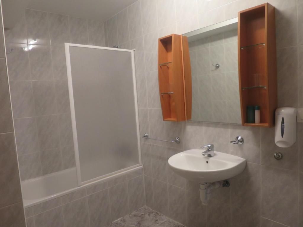 Baño con lavabo blanco, bañera con cristal esmerilado, espejo y muebles de madera.