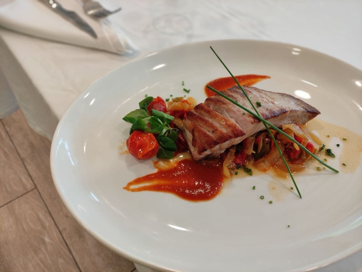 Pescado a la parrilla sobre un lecho de verduras, adornado con cebollino y salsa roja en un plato blanco.