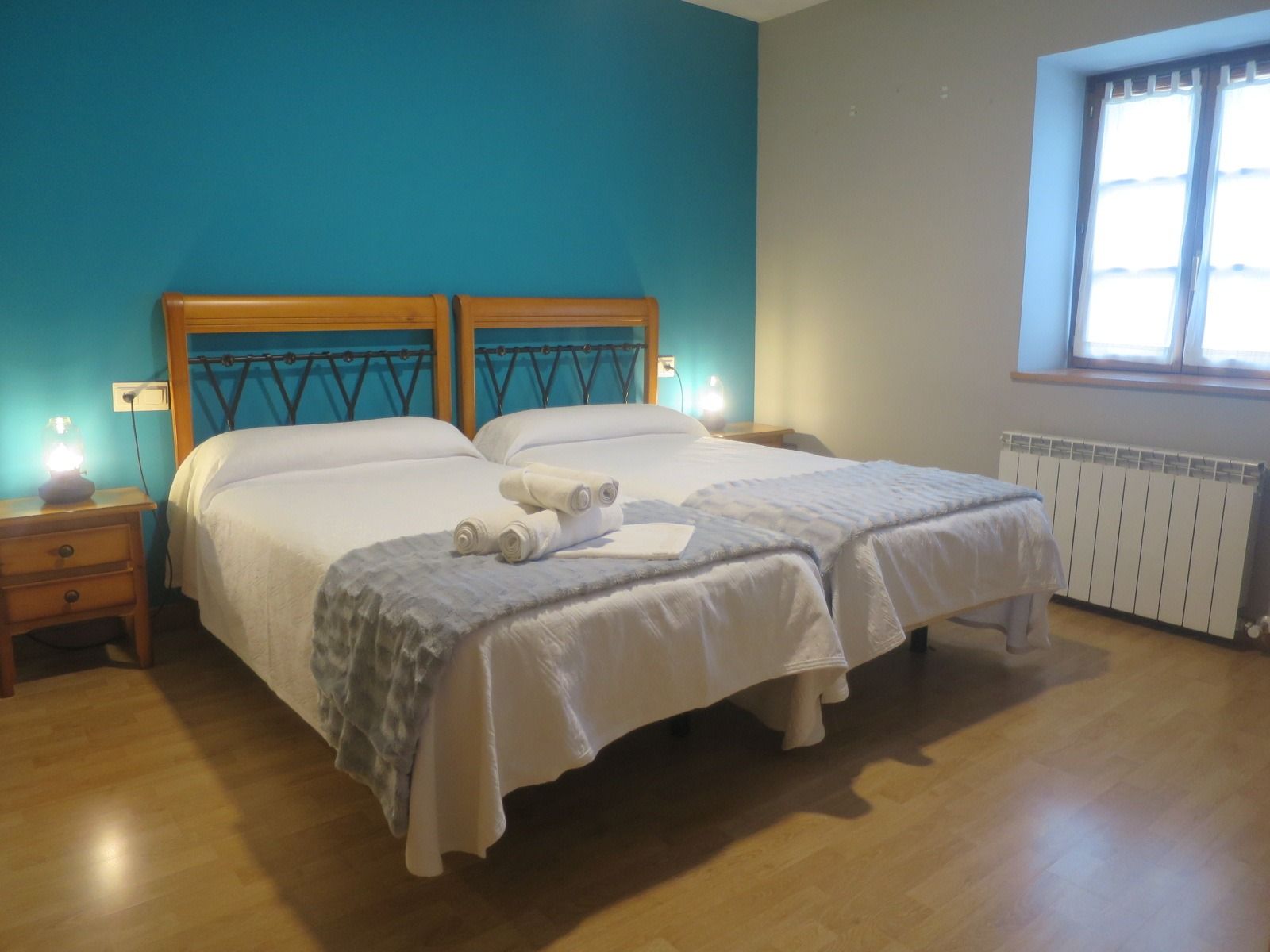 Dormitorio con dos camas individuales, pared con acento verde azulado, mesitas de noche de madera y una ventana.