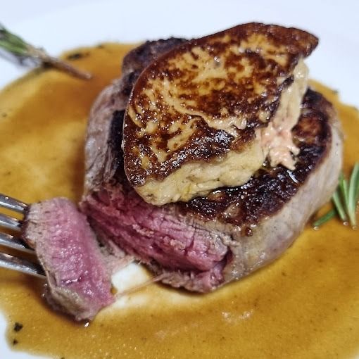 Filete cubierto con foie gras sellado, un bocado que revela un color rosa poco hecho. Salsa marrón, plato blanco, ramitas de romero.