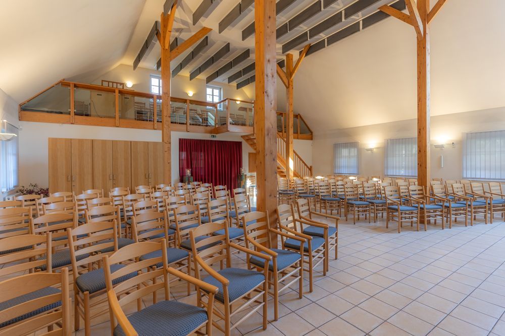 Ein leerer Kirchensaal mit Reihen von Holzstühlen und einer rot verhängten Bühne