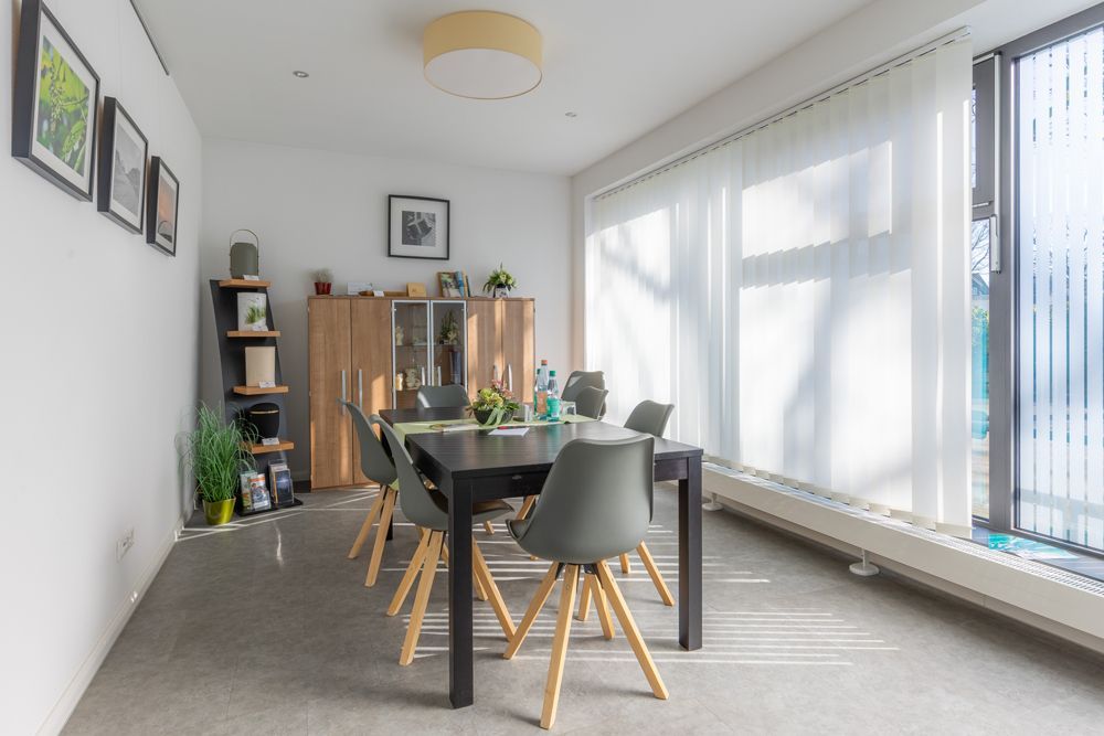 Modernes Esszimmer mit schwarzem Tisch, grauen Stühlen, Pflanzen und großen, sonnendurchfluteten Fenstern